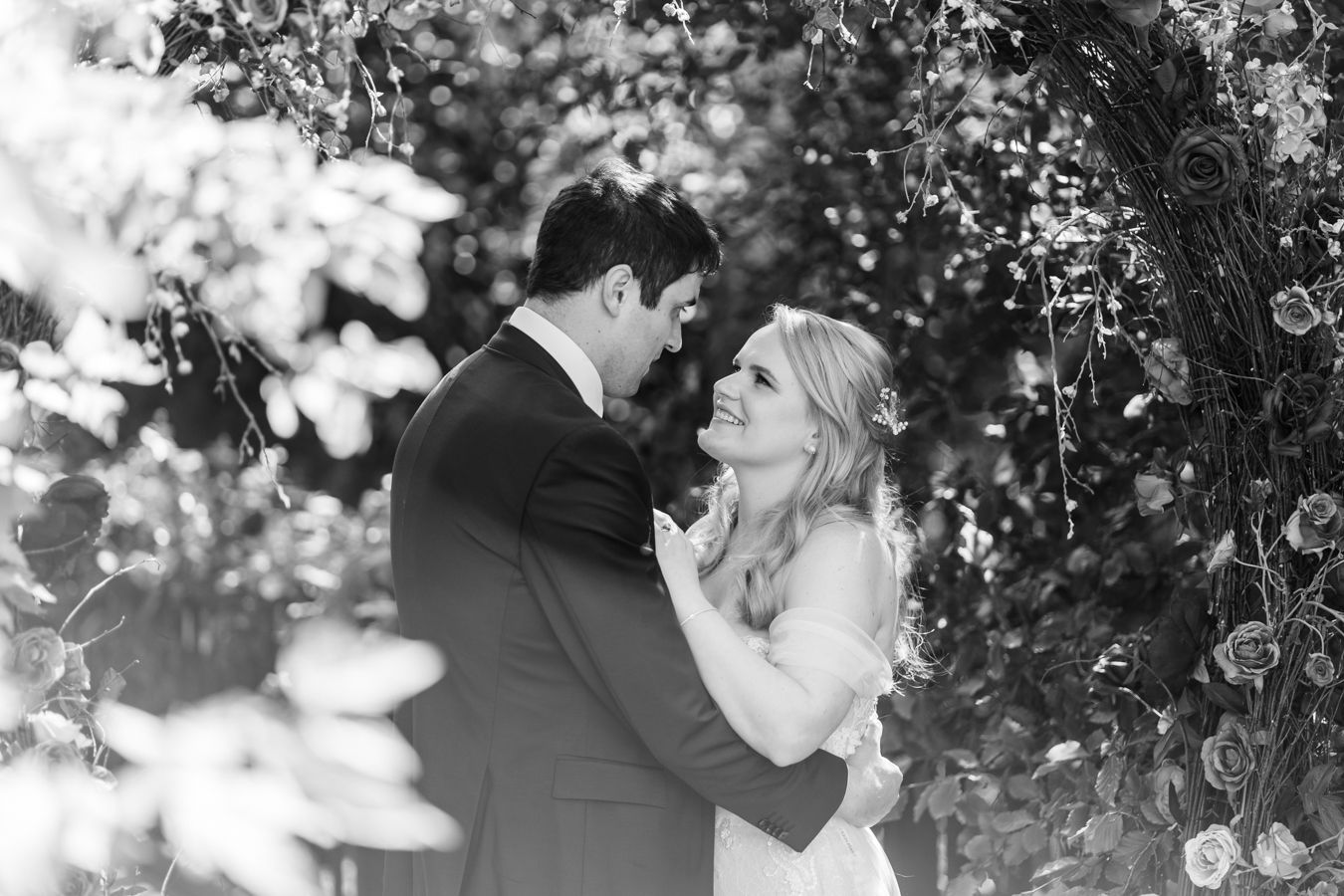 Black and white image of the couple embracing under a rose arch, bride looking up smiling