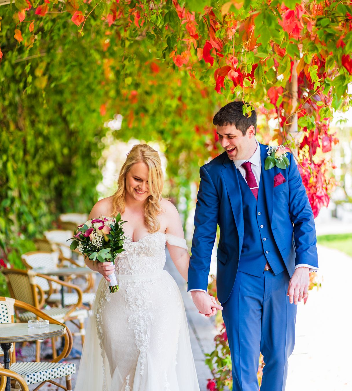 Bride and groom walking hand in hand under a canopy of autumn red ivy, bride holding a small bouquet