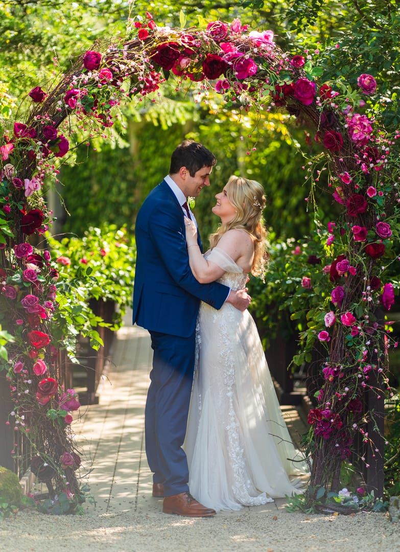 Bride and groom under a rose-covered garden arch in full pink bloom, looking into each other's eyes