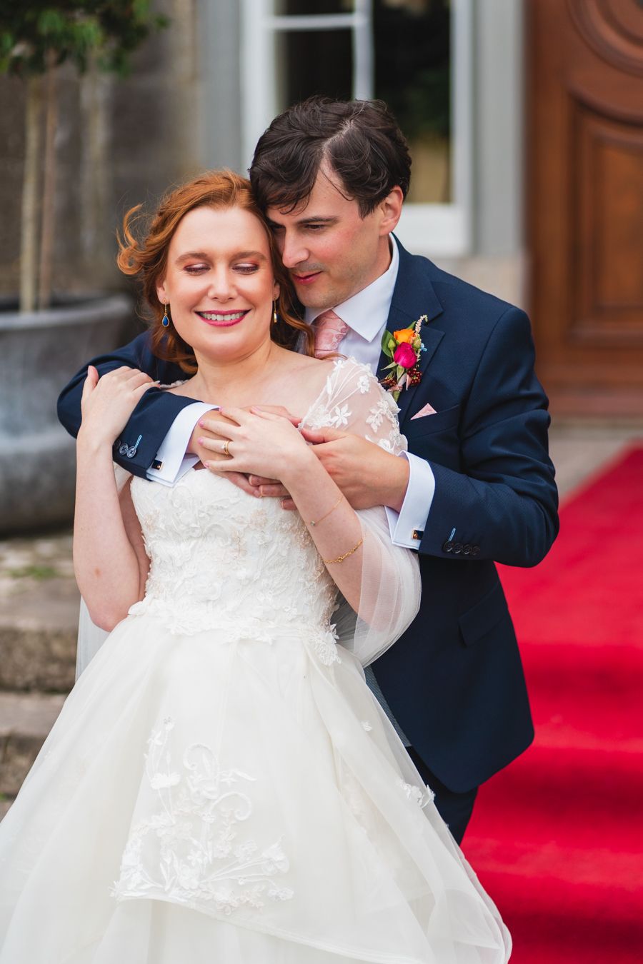 Groom embracing the bride from behind on red-carpet venue steps, both smiling softly