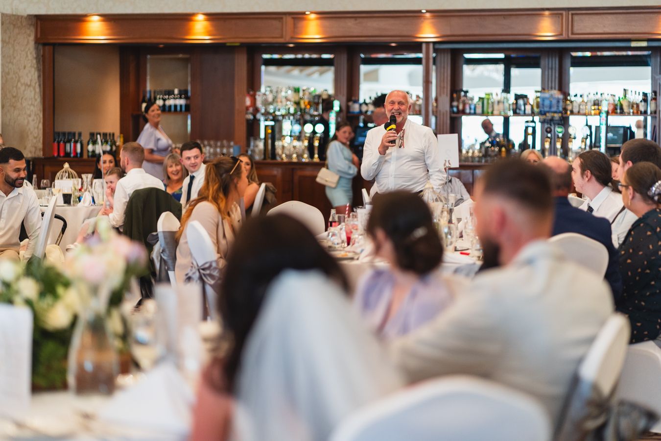 Speaker addressing wedding guests at the reception with a microphone, tables filled with seated attendees