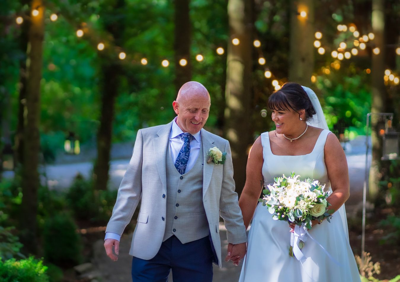 Bride and groom walking through woodland strung with warm fairy lights, bride holding a white bouquet
