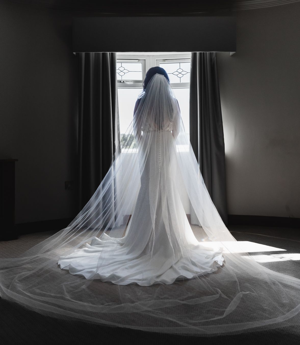Bride alone at a tall window with her long veil pooled across the carpet behind her