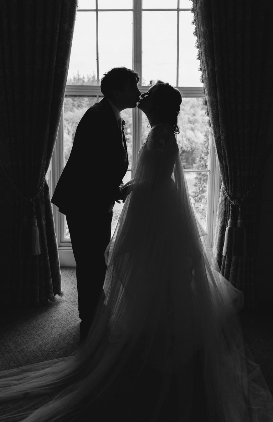 Black and white silhouette of the couple kissing at a tall window, the bride's veil cascading behind her
