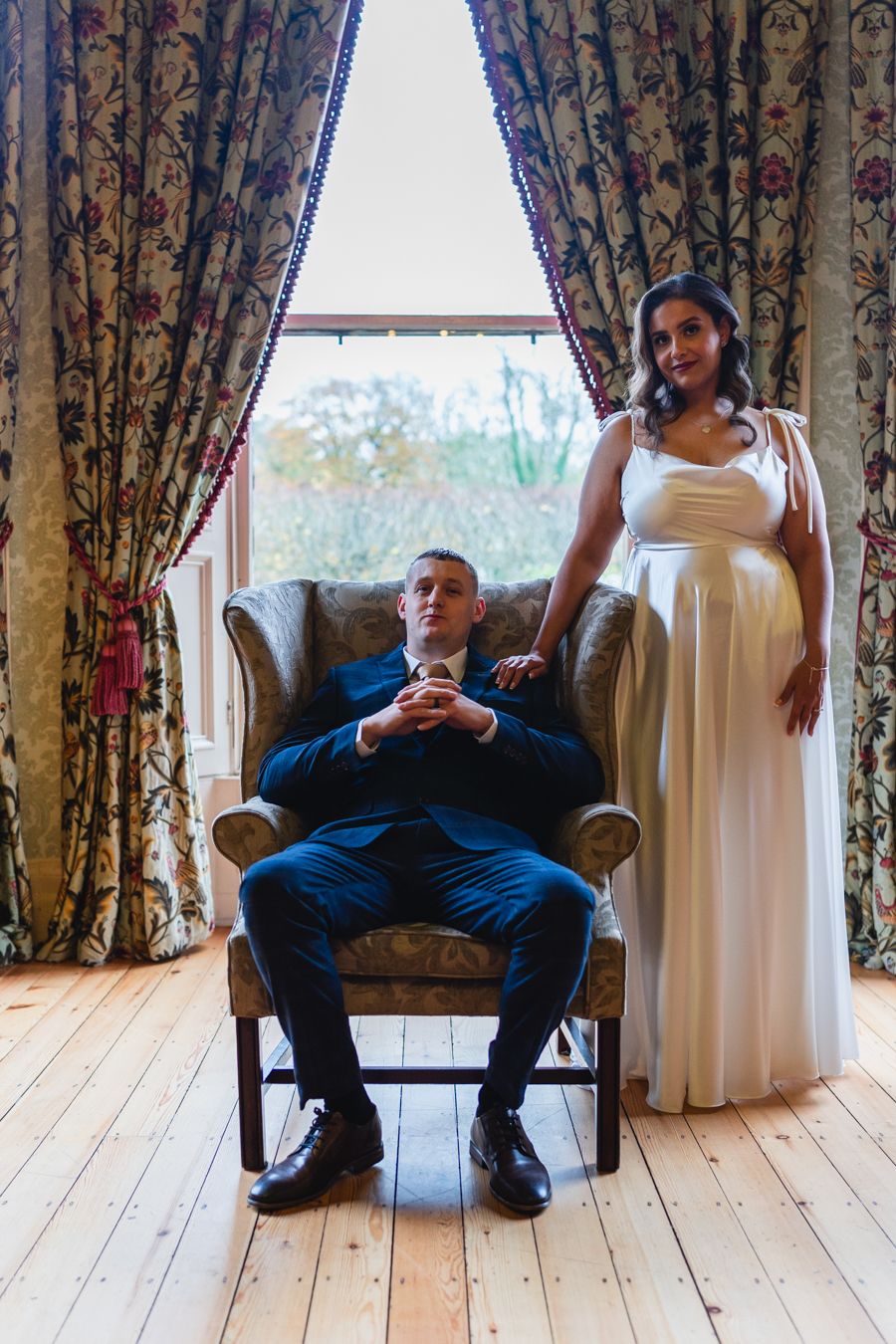 Formal portrait of the groom seated in a patterned armchair with the bride standing beside him at a country house window