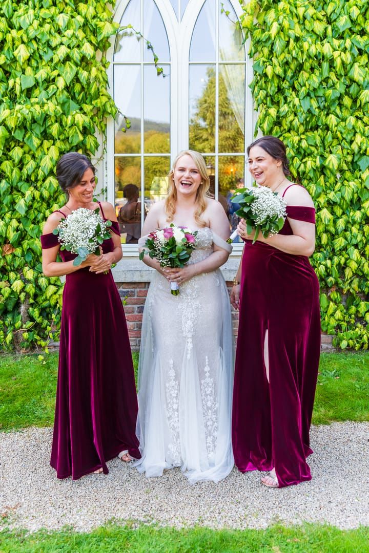 Bride laughing with her two bridesmaids in burgundy dresses in front of an ivy-covered Gothic window