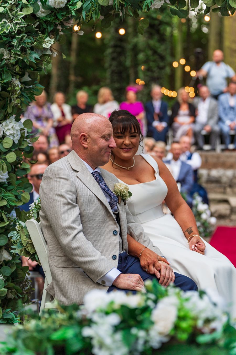 Bride leaning against the groom under a leafy ceremony arch strung with fairy lights, guests seated behind
