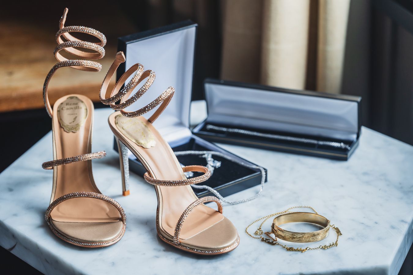 Bridal heels with crystal-coil straps beside a gold bracelet and necklace on a marble surface