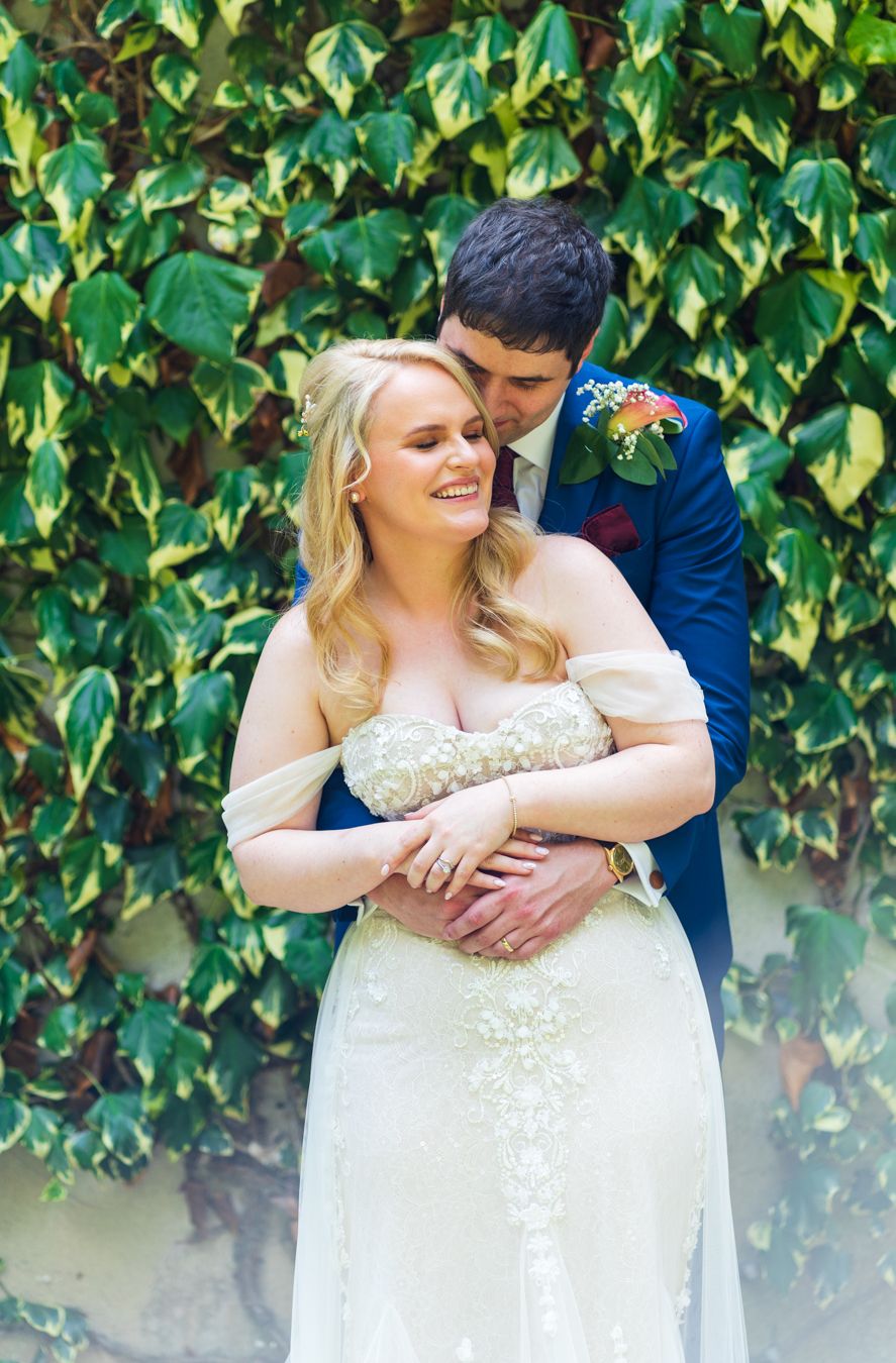 Groom kissing the top of the bride's head against a wall of green ivy